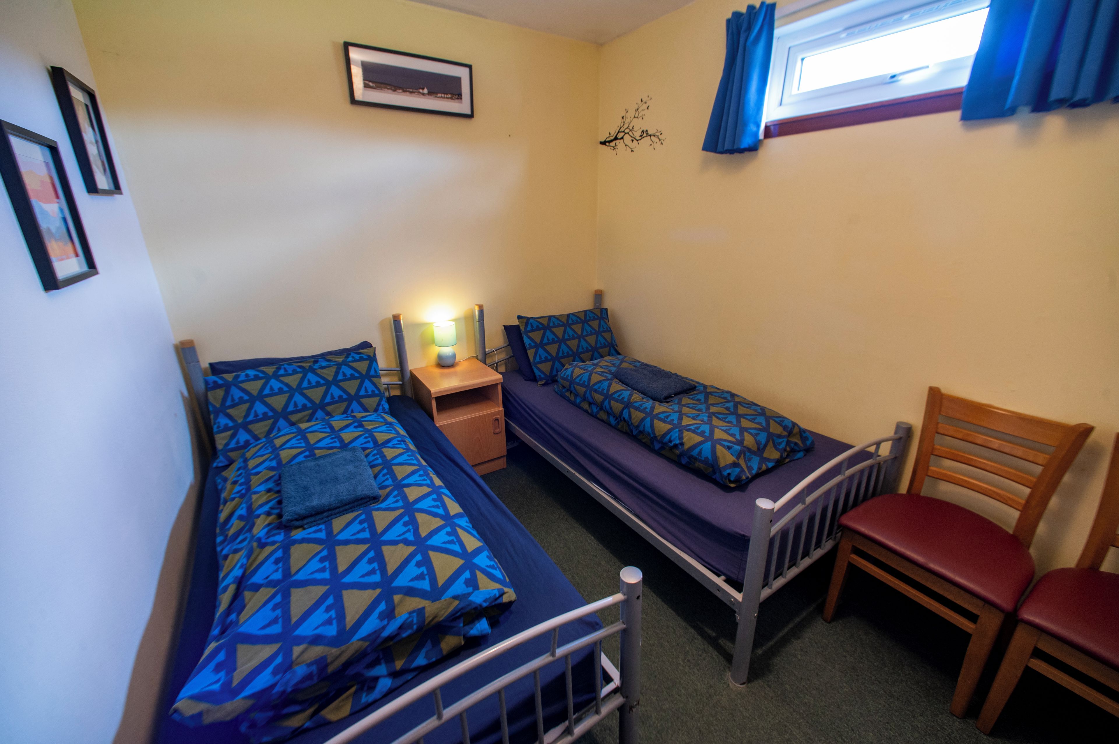 A small, compact room with two single beds, a wooden chair, a nightstand with a lamp, and a window with curtains.