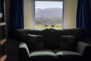 A room with a sofa facing a window overlooking a field with a bench and trees. - Achmelvich Beach Youth Hostel (Achmelvich)