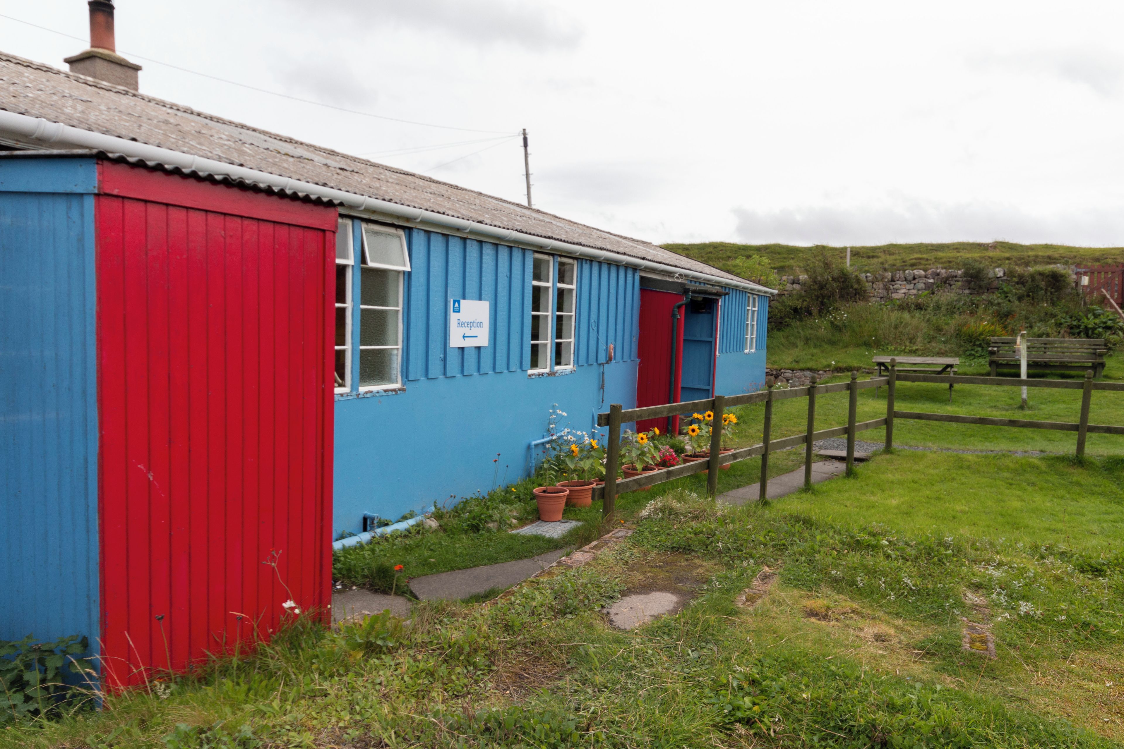 Un petit bâtiment d’un seul étage, avec une porte rouge et des murs bleus, sur lequel on peut voir une enseigne indiquant « réception ».