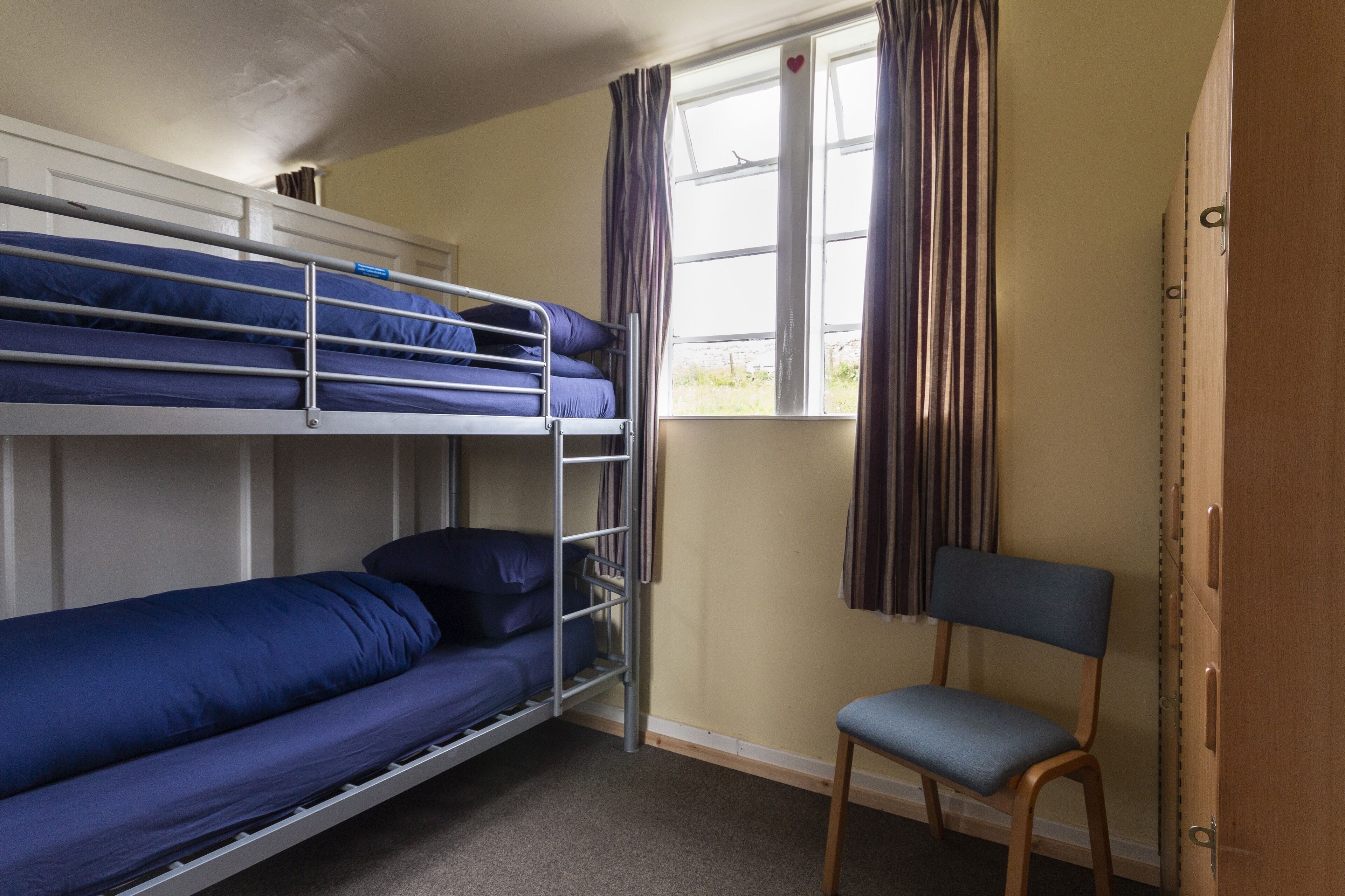 A dormitory room with bunk beds, a chair, and a window with curtains.