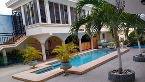 Un'area piscina con una vasca rettangolare, palme e un edificio con numerose finestre e balconi.