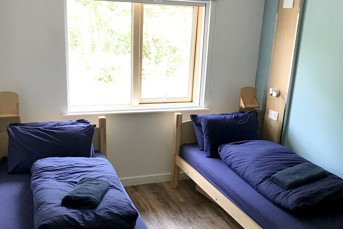 A room with two wooden bunk beds, each with blue bedding and pillows. There is a window providing natural light, and a door with a lock on the right side.