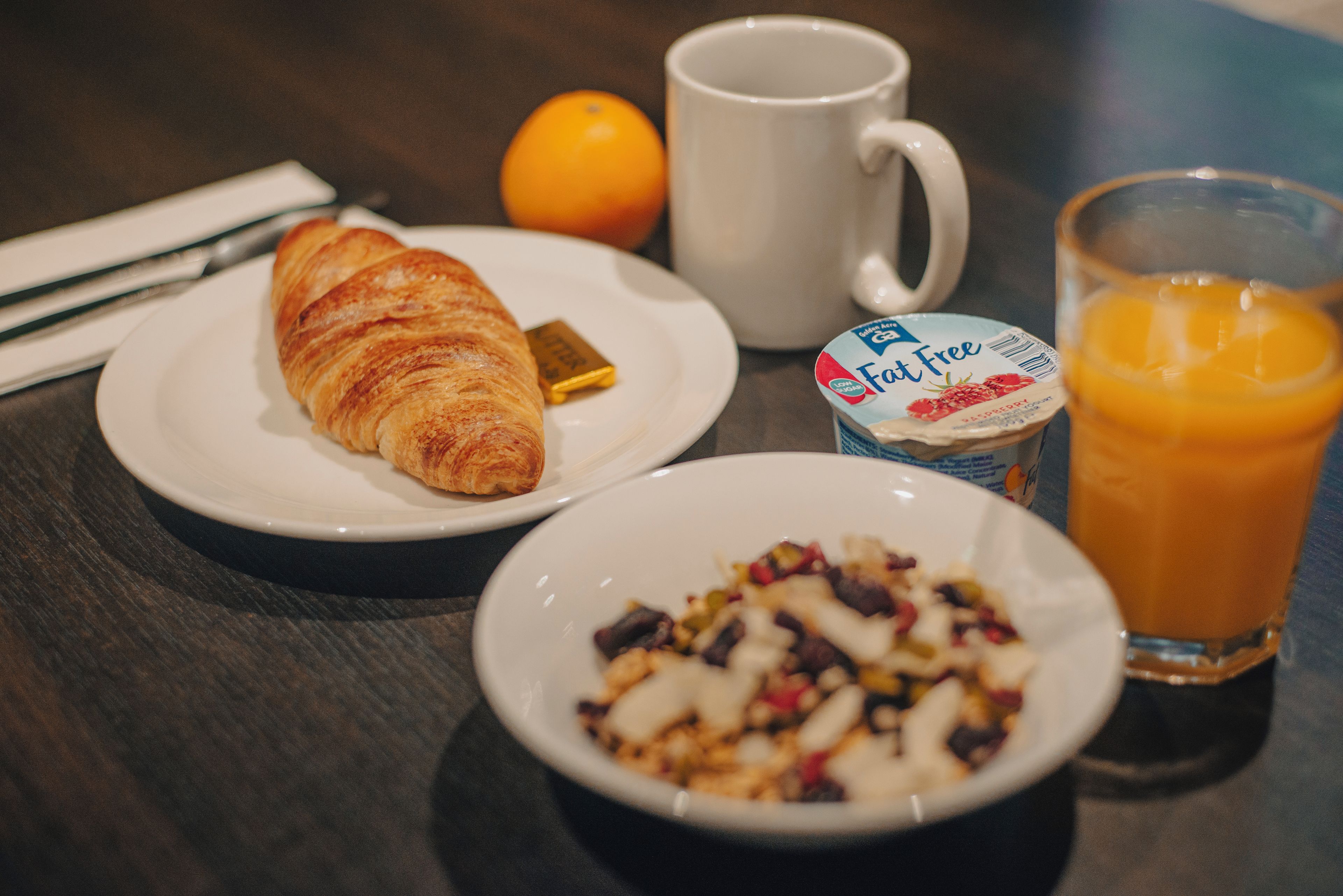 A breakfast setting with a croissant, a glass of orange juice, a mug, and a plate with a yogurt container.