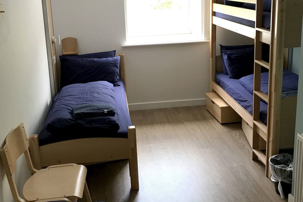 A small, well-lit room with a bunk bed, a single bed, a chair, and a window providing natural light.