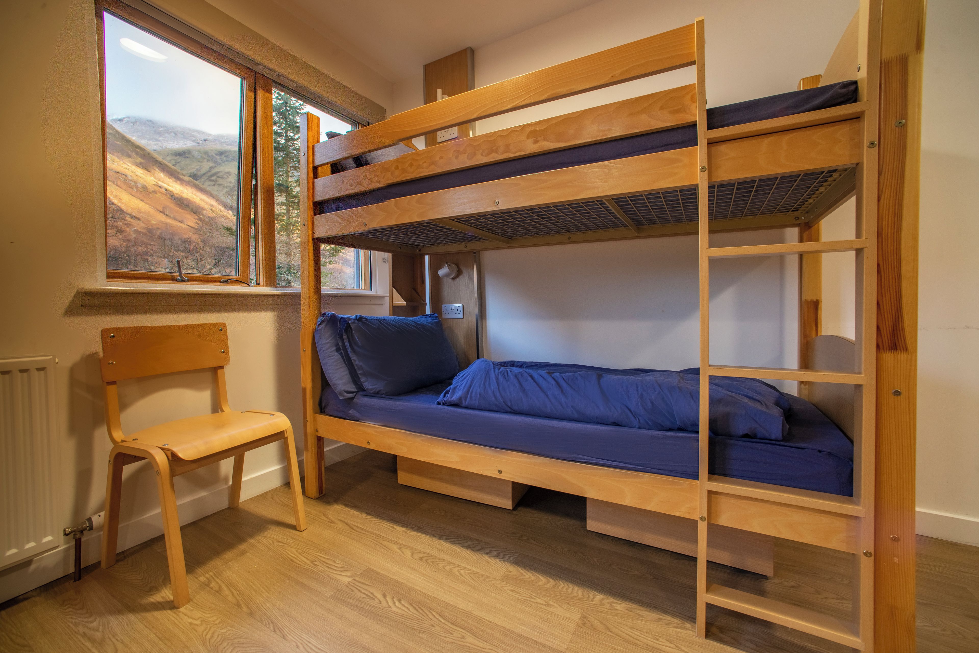 Un lit superposé avec une structure en bois, un lit simple en bas et une chaise à côté.