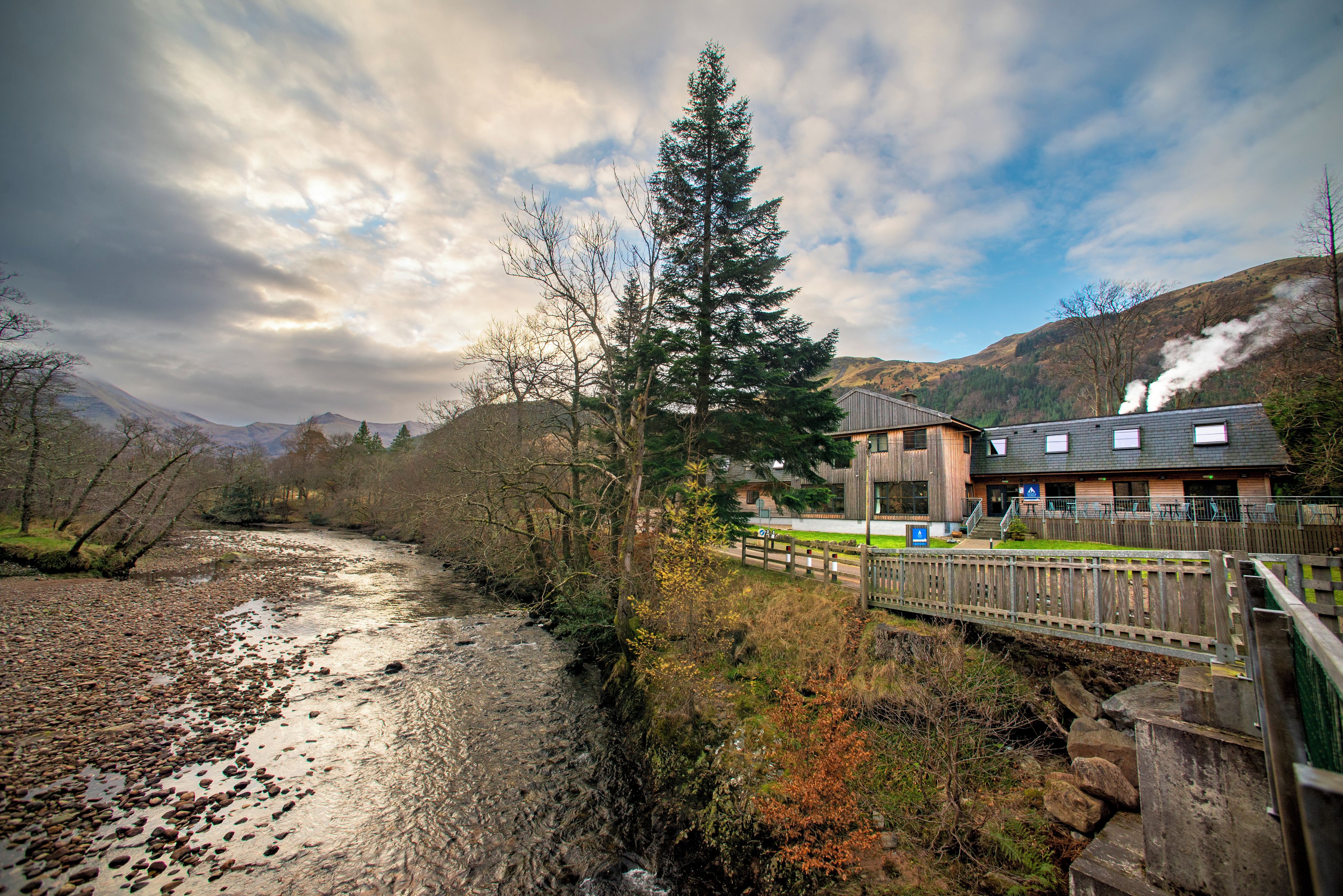 A riverside view with a wooden bridge, a building with a sloped roof, and mountains in the background.