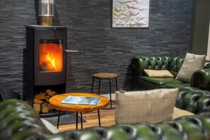 A cozy living room with a stone wall, a green leather sofa, a round wooden coffee table, and a wood-burning stove. - Glen Nevis Youth Hostel (Fort William)