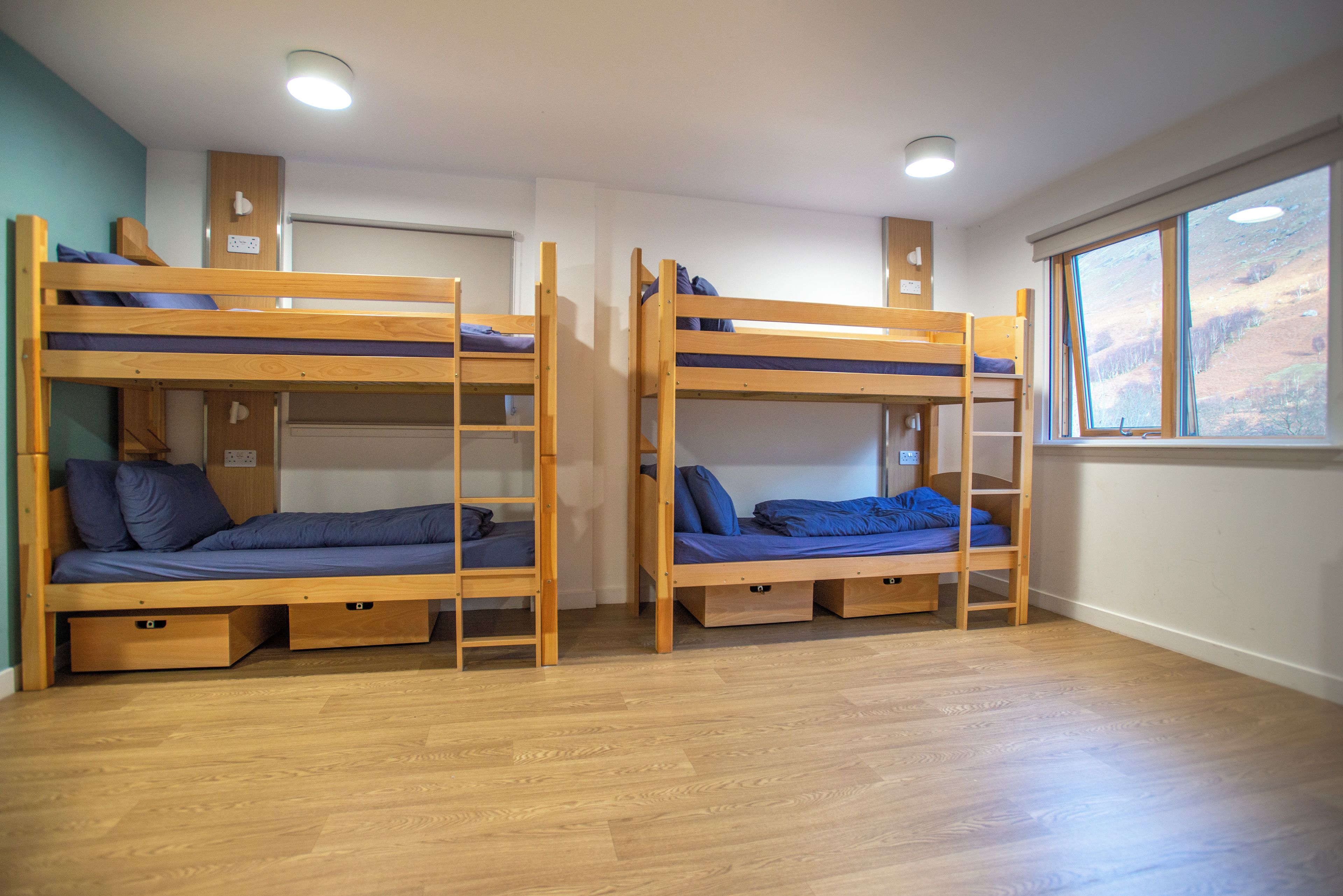 A room with two bunk beds, wooden flooring, a window with a view, and a ceiling light fixture.