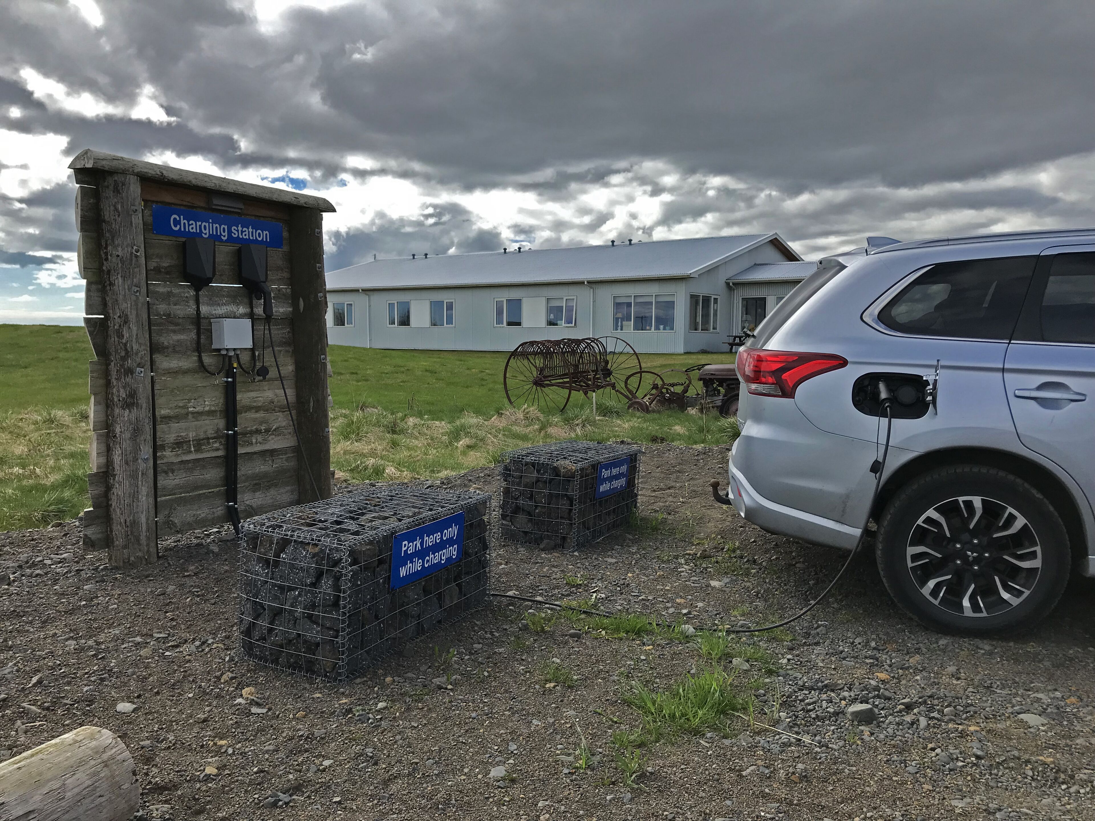Eine Ladestation mit Schild, ein geparktes Auto und ein Gebäude im Hintergrund.