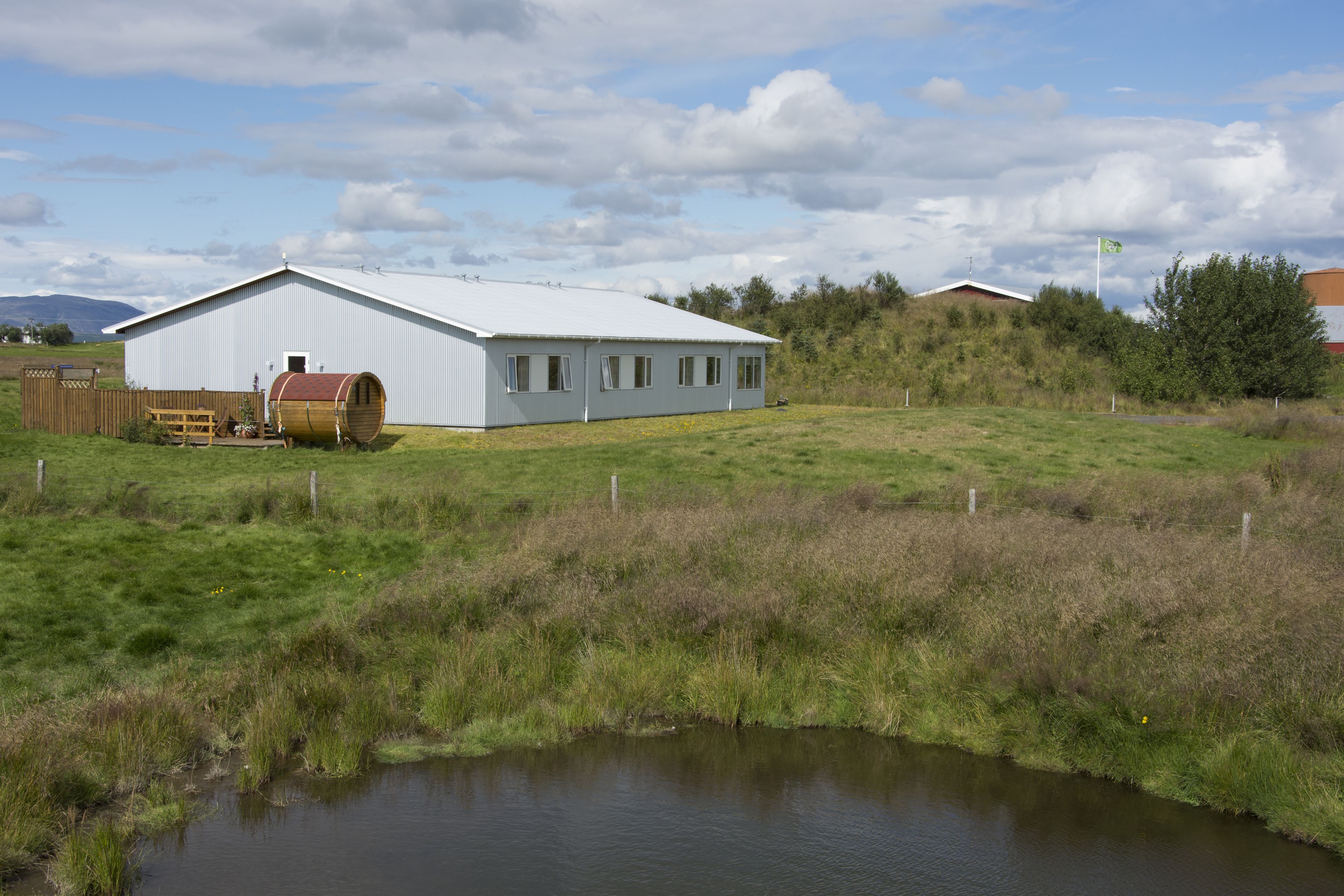 A large white building with a flat roof, a wooden fence, and a small pond in front.