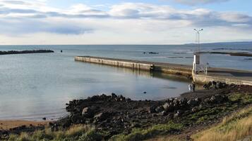 Un paysage côtier avec une rive rocheuse, une jetée en bois s’avançant dans des eaux calmes et un phare situé à droite.