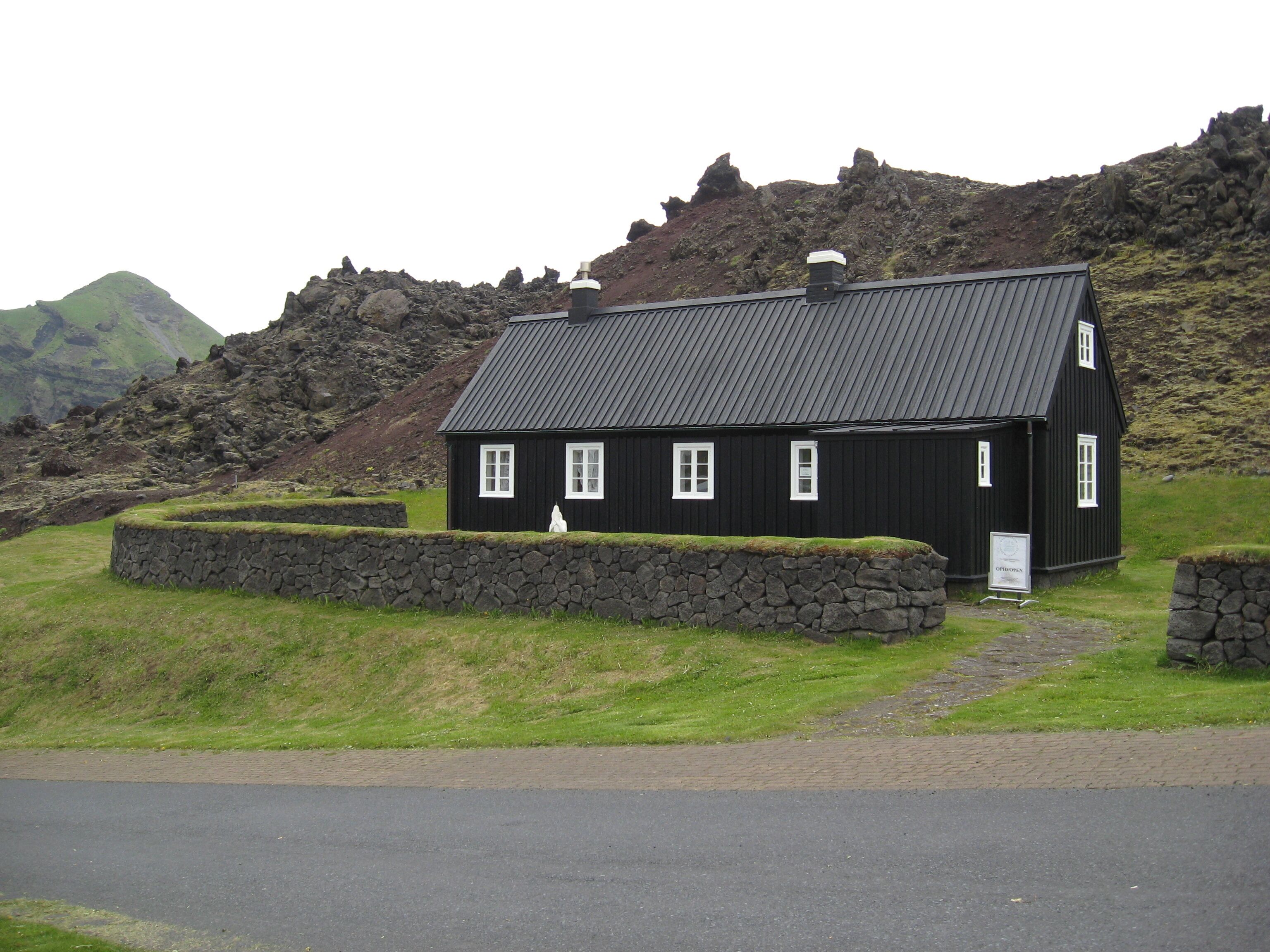 Una casa de madera negra con un muro de piedra y un camino de piedras que lleva a ella, con montañas al fondo.