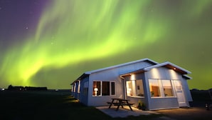 A modern house with large windows and a patio, under a night sky with visible aurora borealis. - Lambastadir Guesthouse (Selfoss)