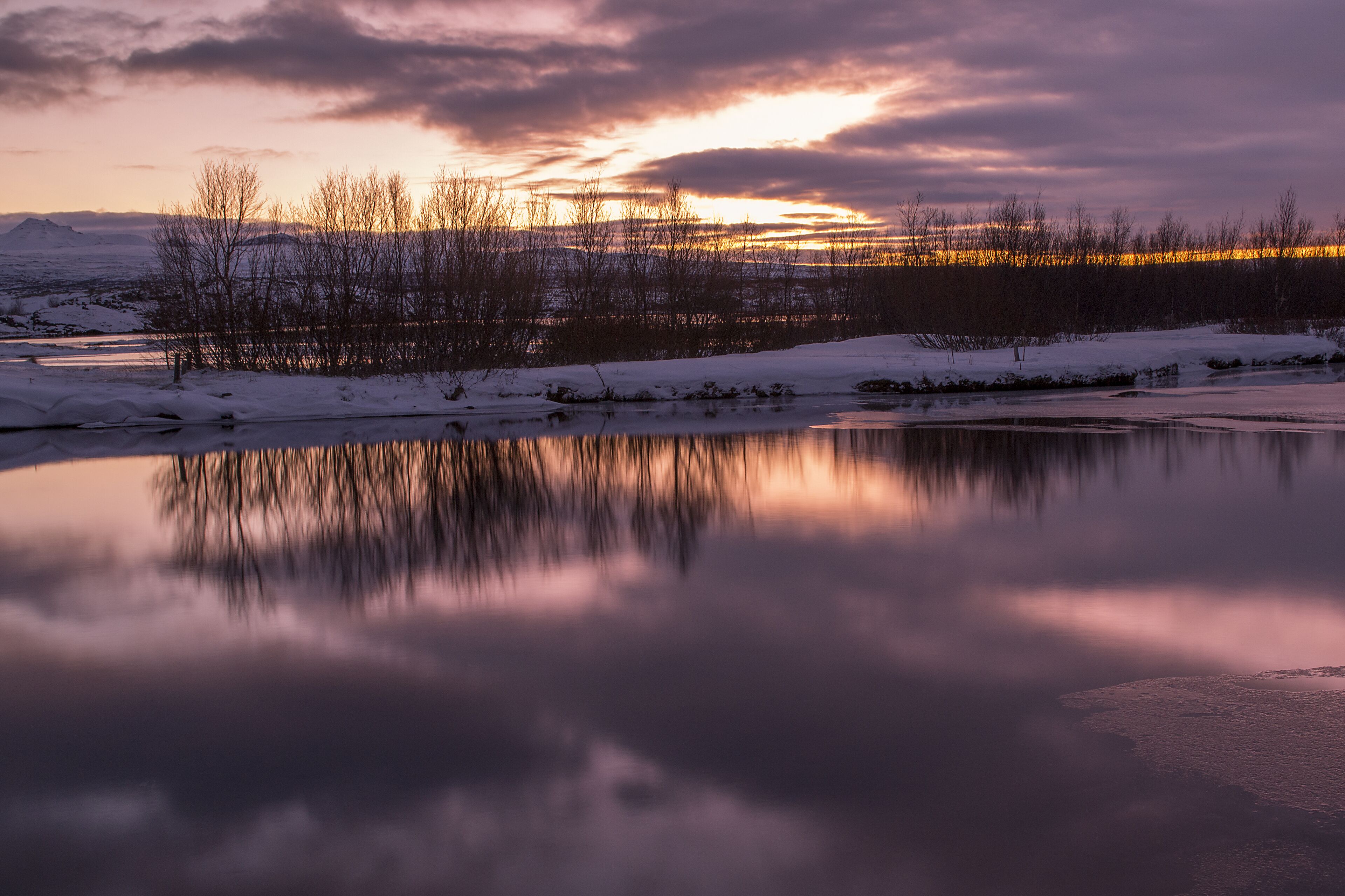 Un paisaje sereno con un lago congelado que refleja la puesta de sol, rodeado de terreno cubierto de nieve y árboles sin hojas.