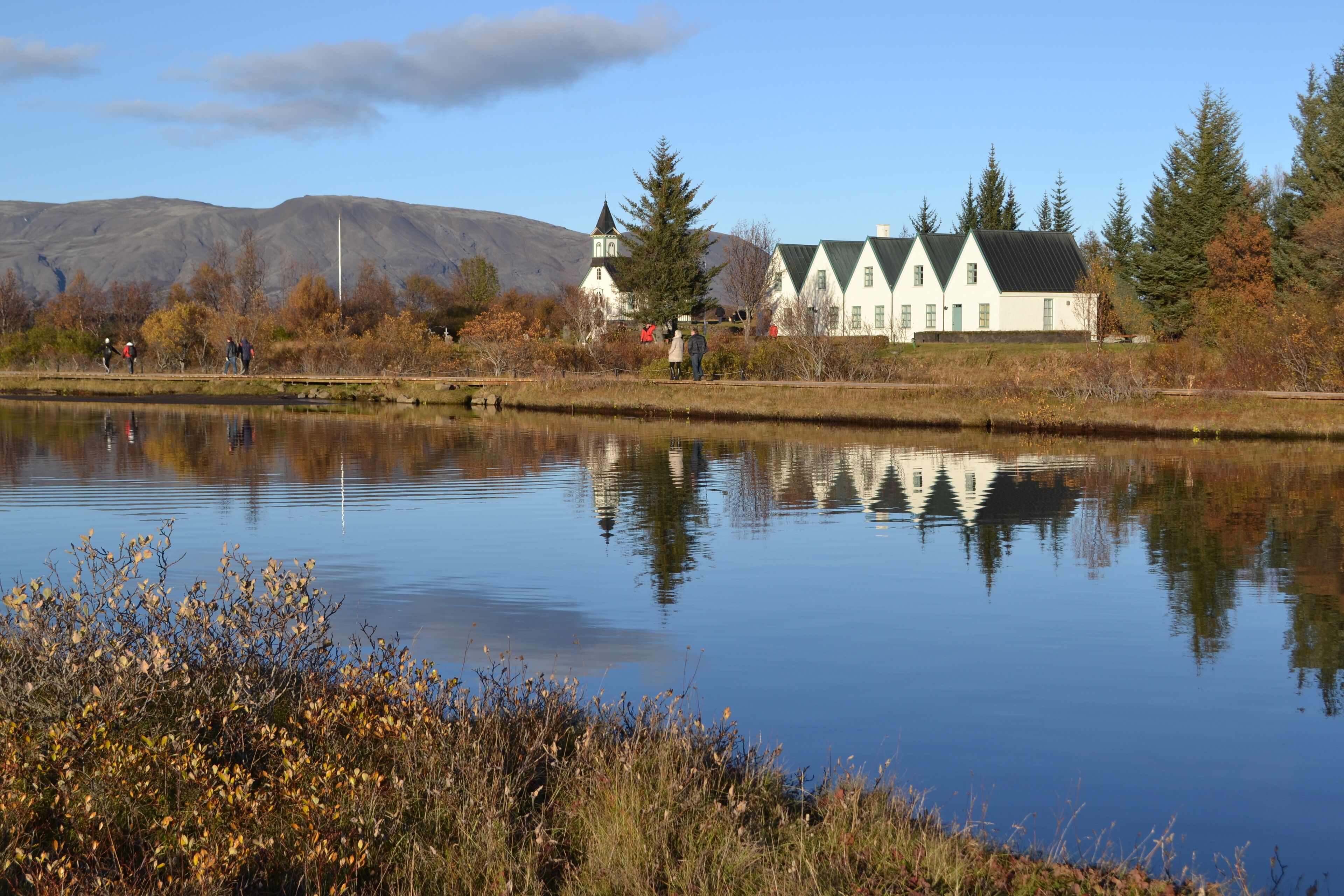A serene lakeside setting with a white building featuring a traditional architectural style, surrounded by autumn foliage and a calm water body reflecting the structure.