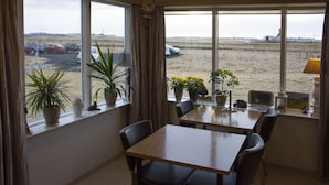 A dining area with a table and chairs, potted plants, and a view of a parking lot and beach through large windows. - Lambastadir Guesthouse (Selfoss)