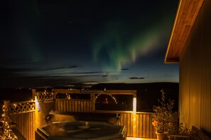 A hot tub area with a wooden deck, a covered seating area, and a view of the northern lights. - Lambastadir Guesthouse (Selfoss)
