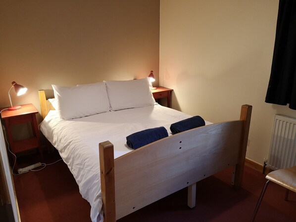 Una cama individual con estructura de madera, ropa de cama blanca, dos mesitas de noche con lámparas y un radiador en la pared.