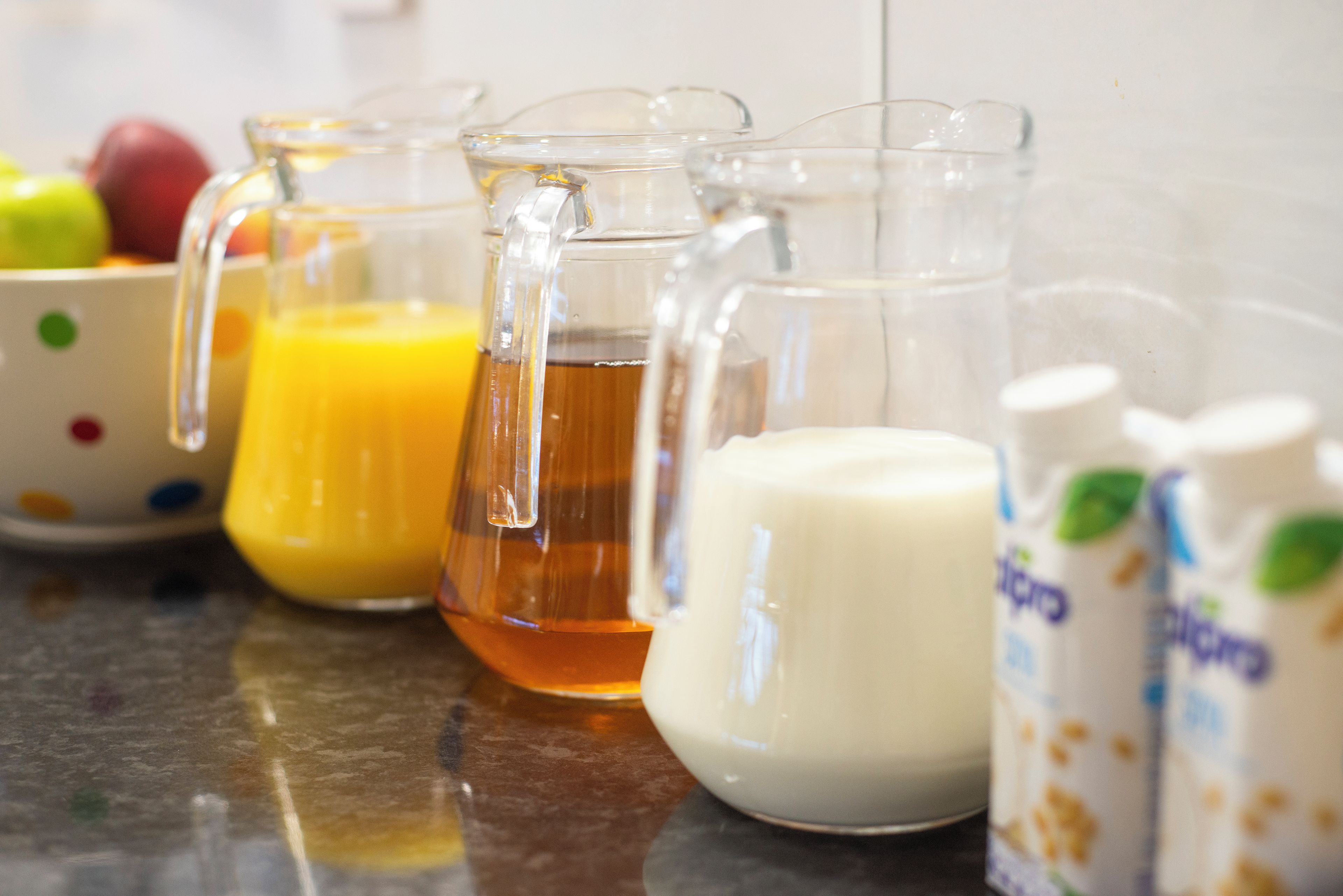 A kitchen counter with various glass containers holding different liquids, a white bowl, and a polka-dotted plate with fruits.