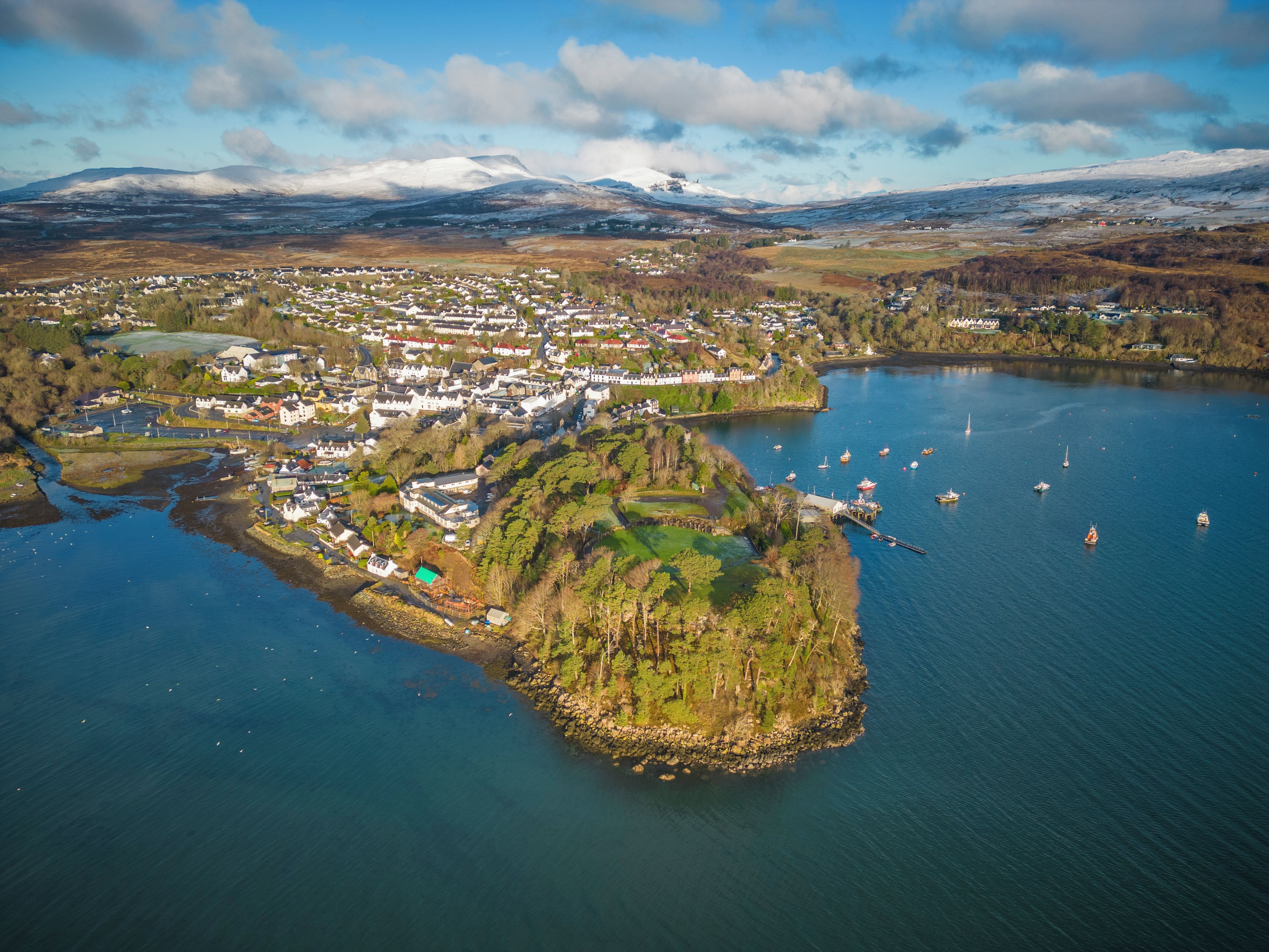 An aerial view of a coastal town with a harbor, boats, and a mountainous backdrop.