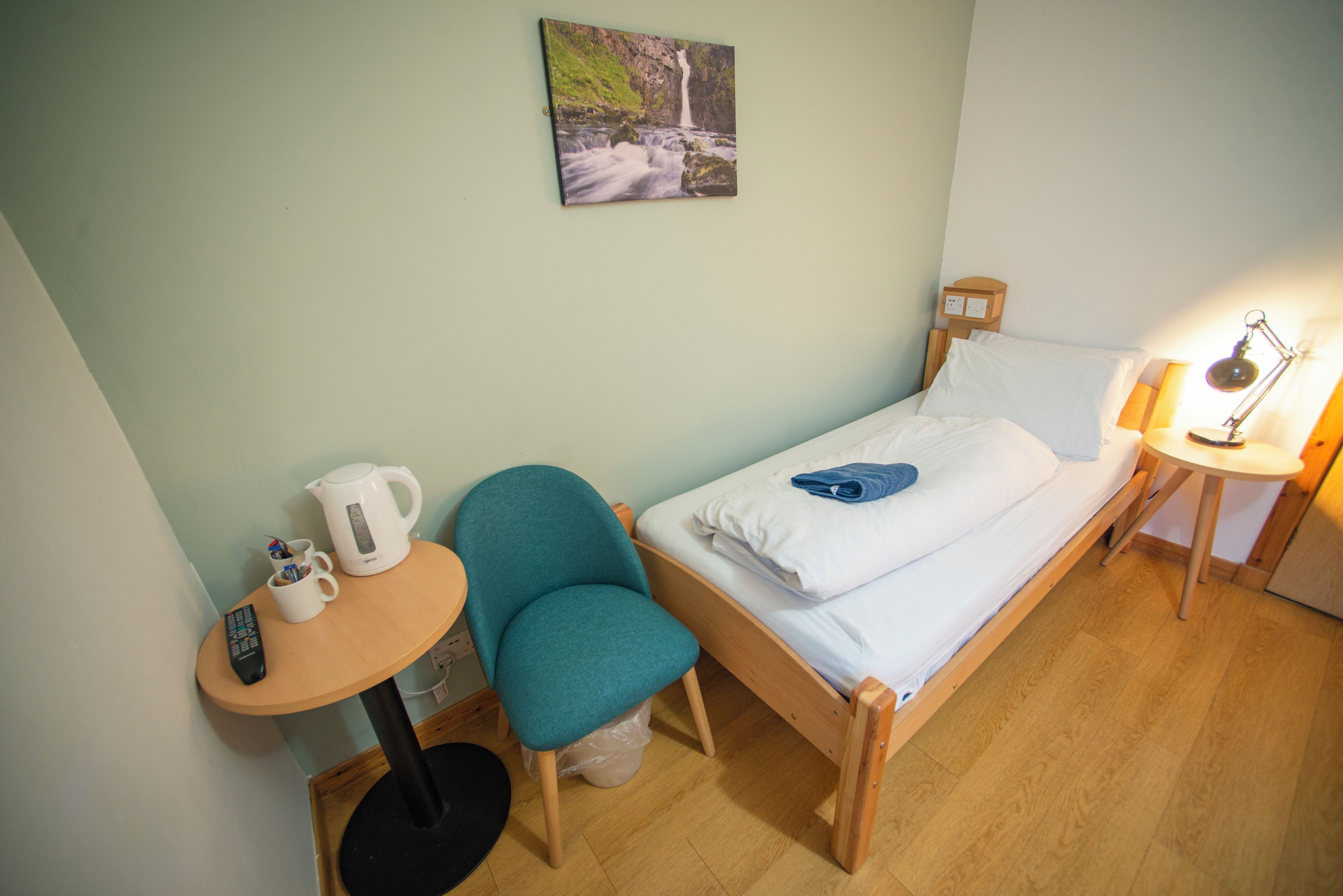 A small, tidy room with a single bed, a chair, a small table, a kettle, and a lamp.