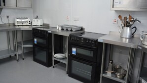 Eine gewerbliche Küche mit Edelstahlgeräten, darunter zwei Öfen und ein Toaster, sowie einem Metallschränkchen mit der Aufschrift 'Gläser'.