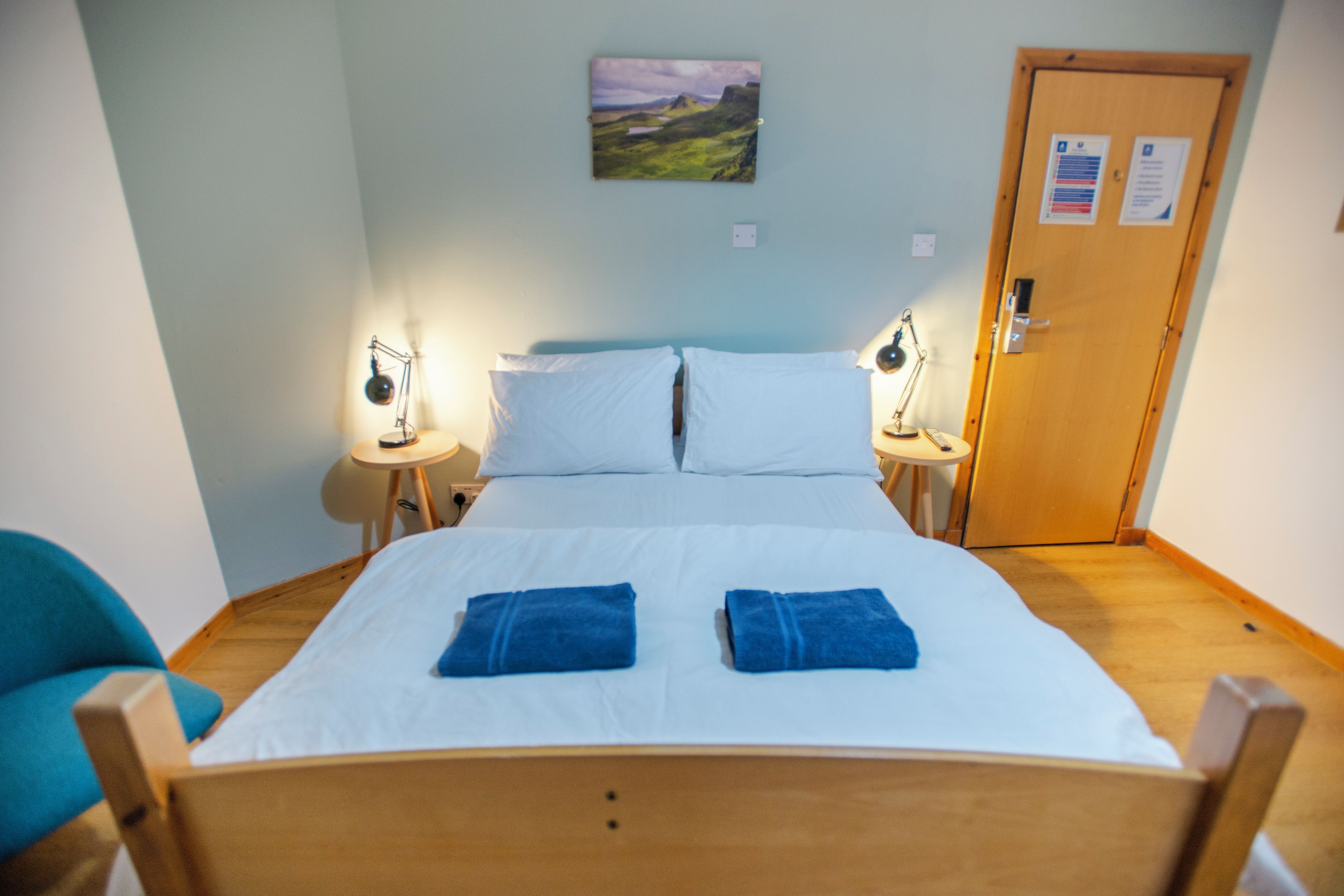 A single-bed room with a wooden headboard, two bedside tables with lamps, a door with a notice, and a landscape painting on the wall.
