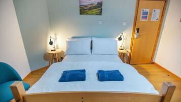 A single-bed room with a wooden headboard, two bedside tables with lamps, a door with a notice, and a landscape painting on the wall.
