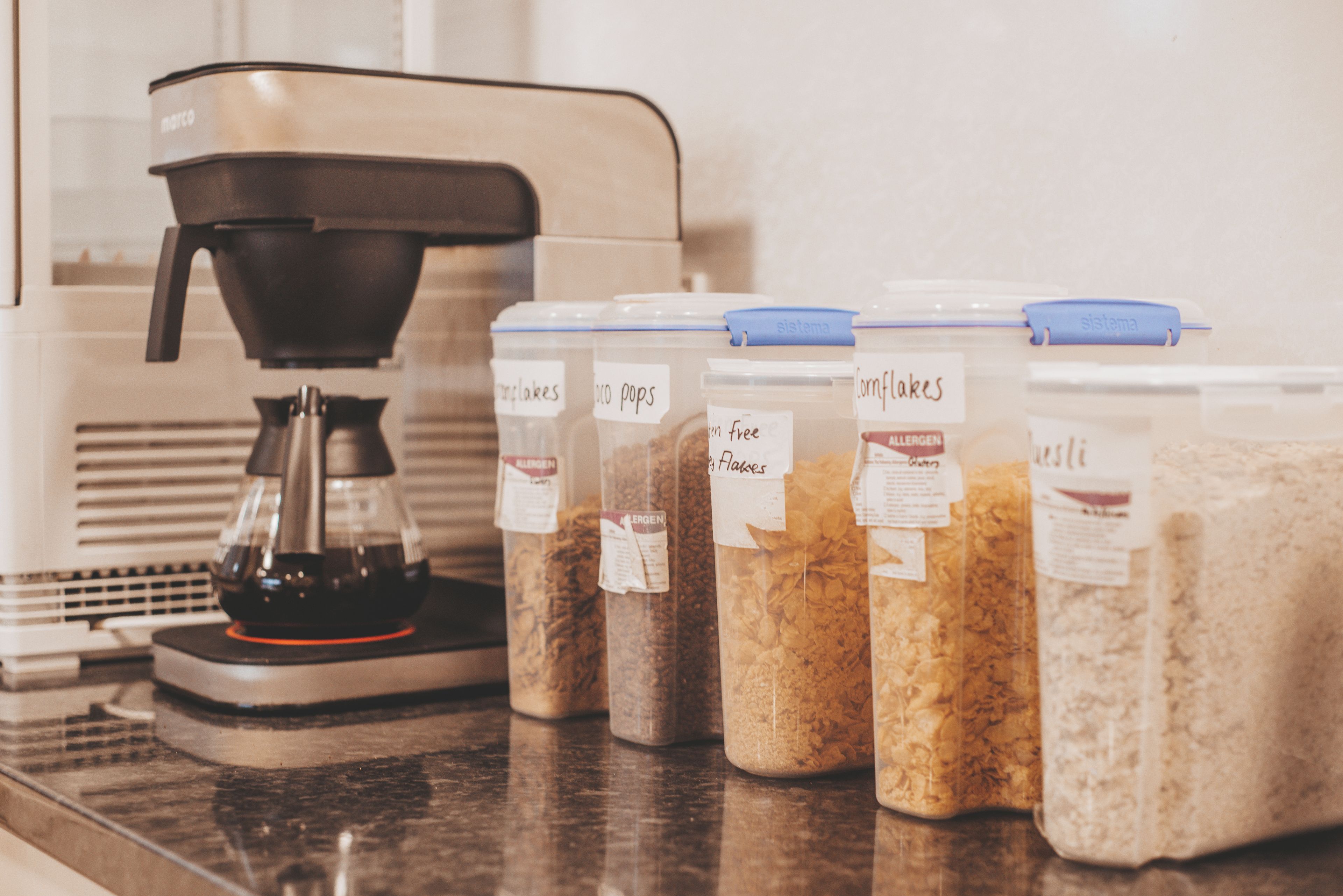 Una encimera de cocina con una cafetera y recipientes etiquetados de cereales y harinas.