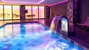 Piscina interior moderna con caída de agua, rodeada de sillones de descanso y una zona cerrada con vidrio y asientos.