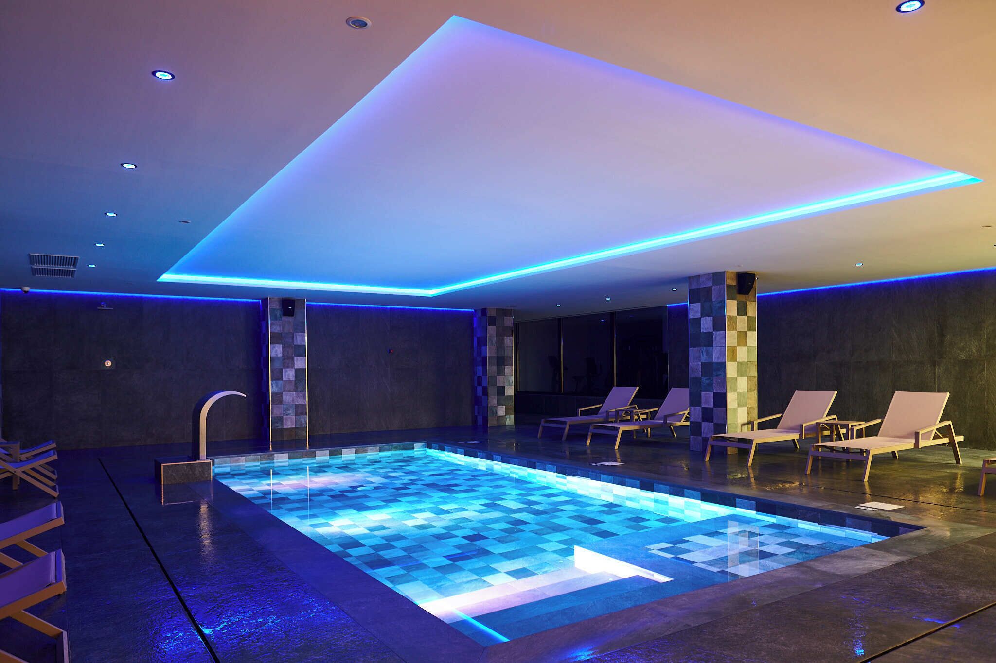 Piscina interior moderna com iluminação azul, espreguiçadeiras e padrão de piso quadriculado.