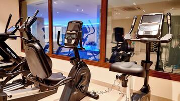 Ein Fitnessraum mit Ergometer und Laufband, ein großes Fenster und ein Spiegelbild eines Schwimmbeckens.