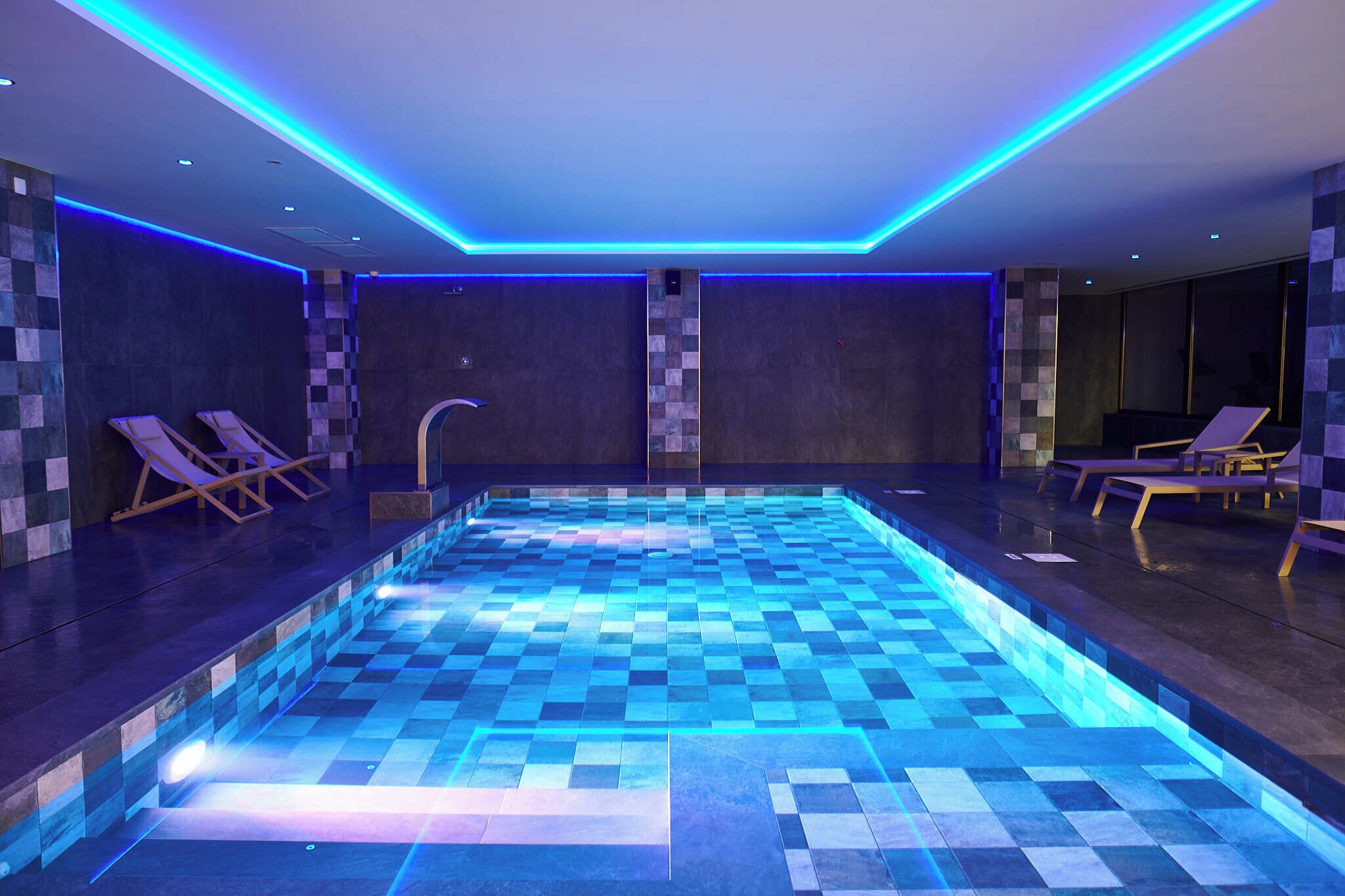 A modern indoor swimming pool with blue lighting, lounge chairs, and a checkered floor pattern.