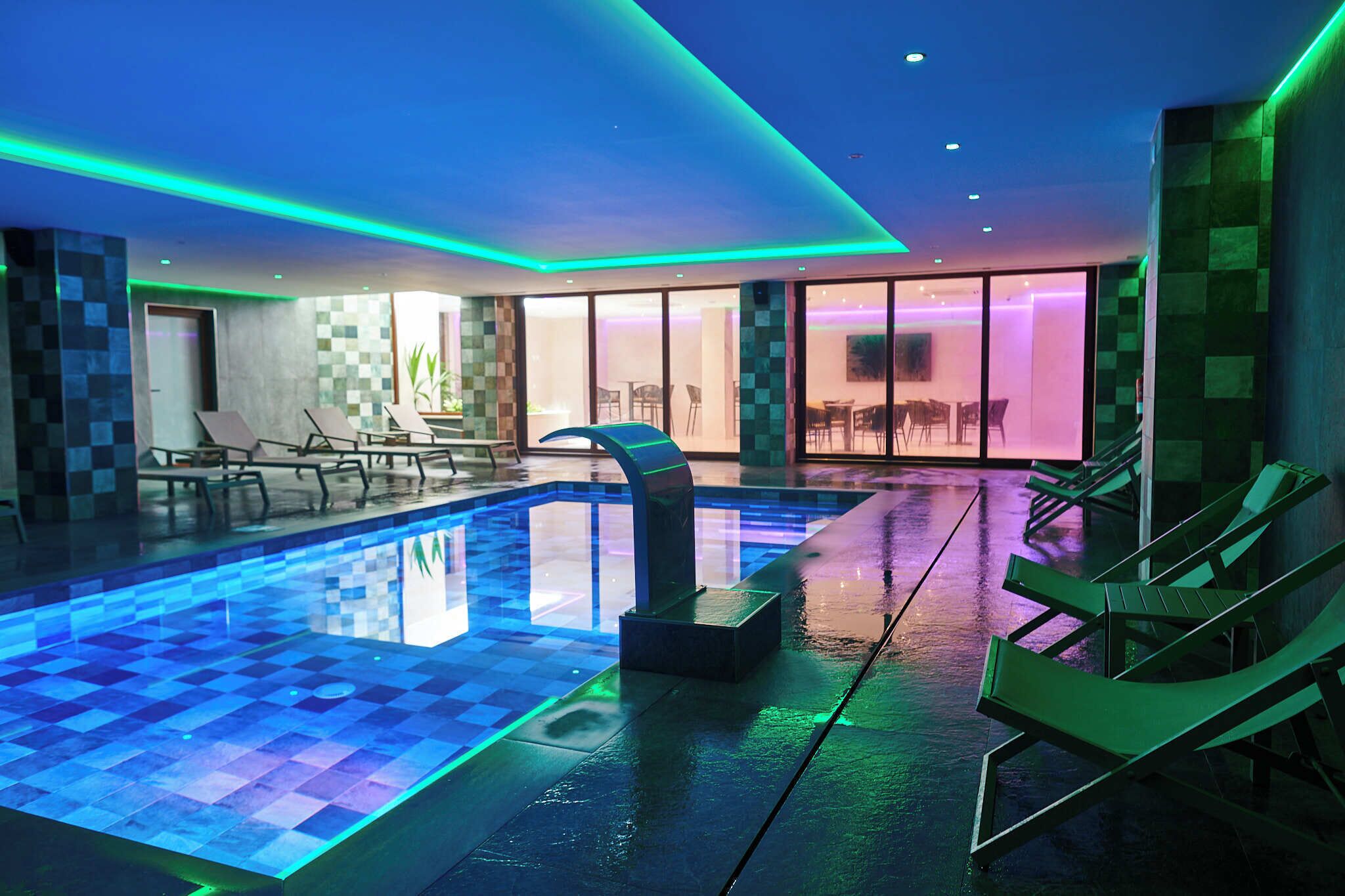 Una piscina interior con diseño moderno, que incluye una estructura curva distintiva en el centro, rodeada de sillones de descanso y un muro de cristal que conduce a un comedor.