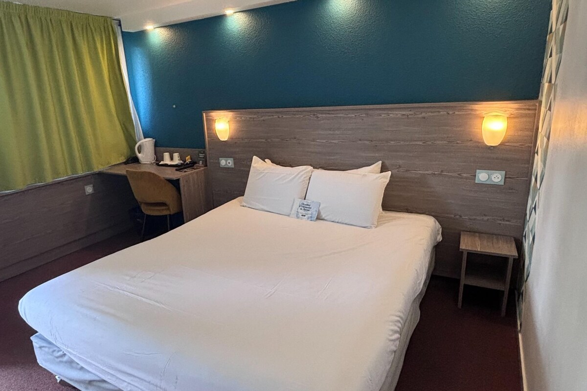 A hotel room with a large bed, a desk with a chair, a bedside table, a fan, and a green curtain.