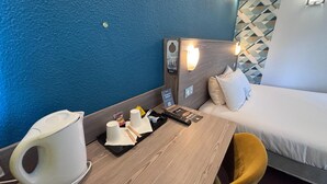 A hotel room with a bed, a wooden table, a white chair, a coffee maker, a tray with cups and snacks, a remote control, and a wall-mounted light. - Kyriad Cambrai (Fontaine-Notre-Dame)