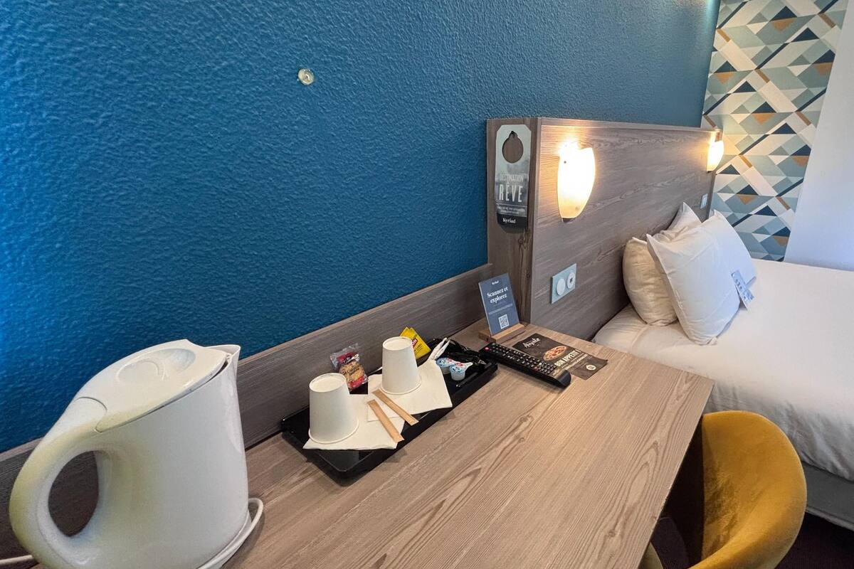 A hotel room with a bed, a wooden table, a white chair, a coffee maker, a tray with cups and snacks, a remote control, and a wall-mounted light.