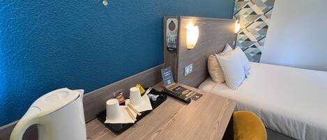 Habitación de hotel con cama, mesa de madera, silla blanca, cafetera, bandeja con tazas y aperitivos, control remoto y una lámpara fijada a la pared.