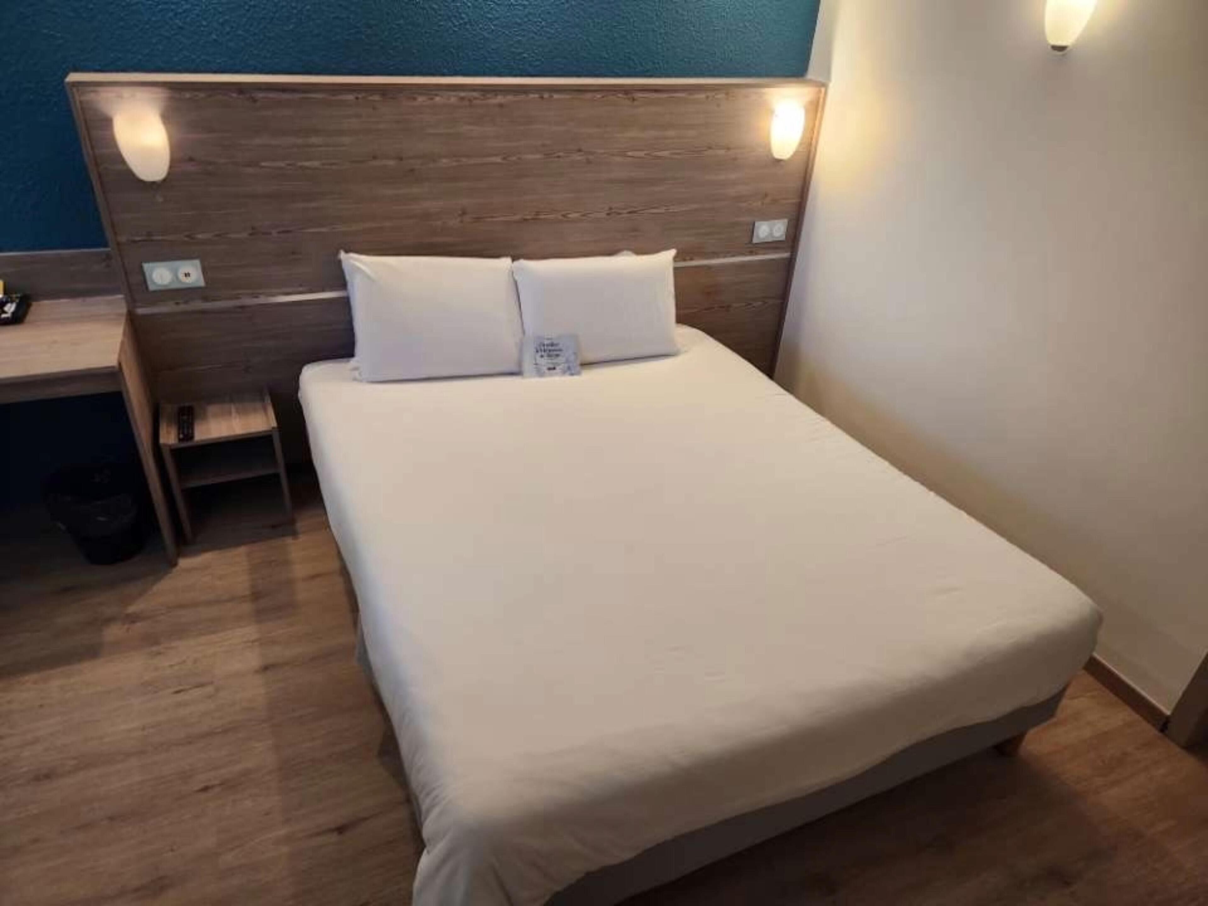 Un lit bien fait, avec du linge de lit blanc, une tête de lit en bois et une petite table de chevet sur laquelle se trouve une lampe.