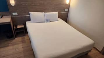 Un lit bien fait, avec du linge de lit blanc, une tête de lit en bois et une petite table de chevet sur laquelle se trouve une lampe.