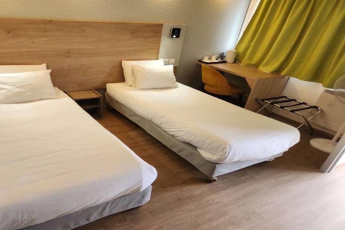 A hotel room with two single beds, a wooden headboard, a small desk with a chair, and a window with curtains.