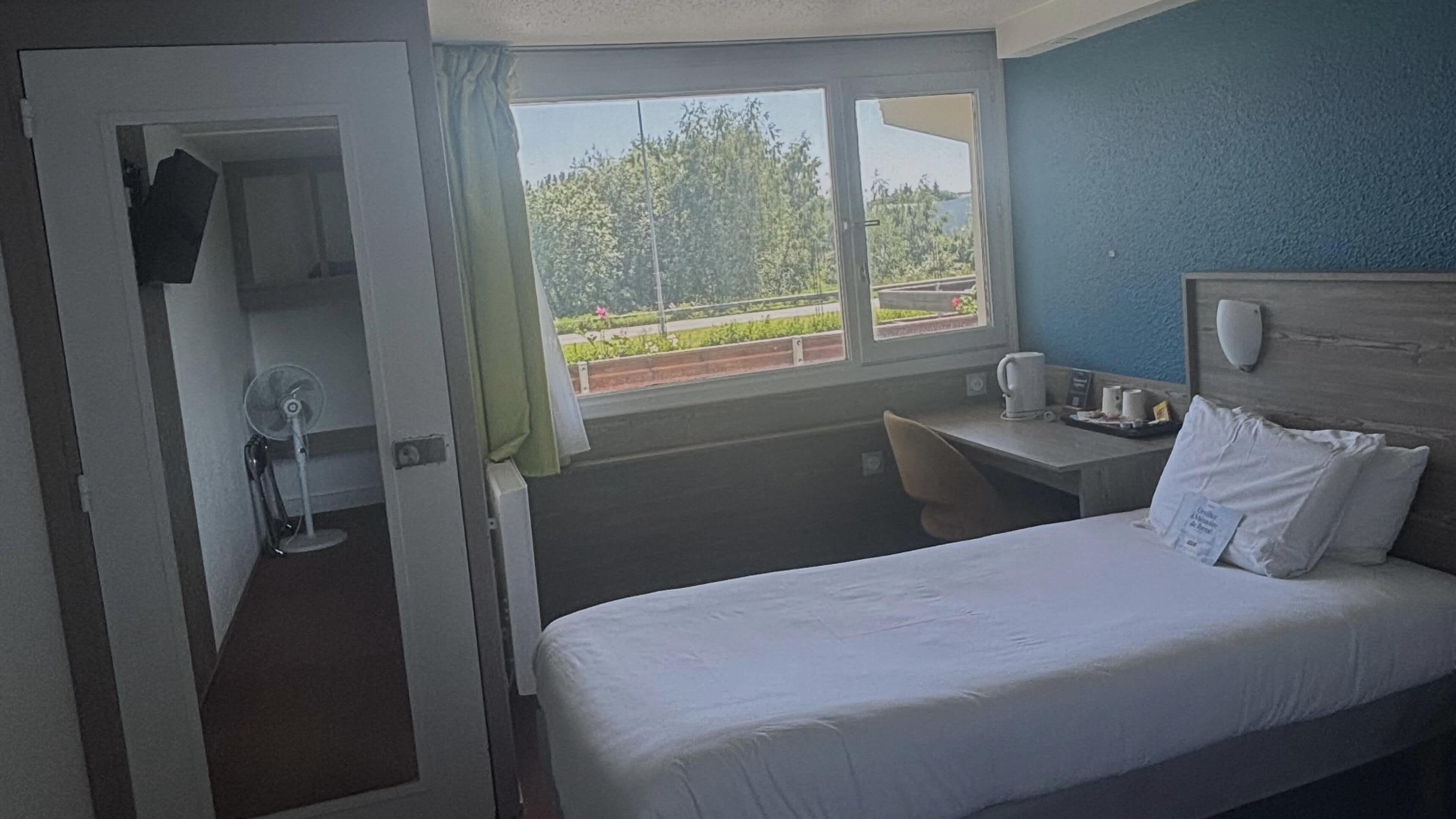 Una camera d'albergo con un letto, una scrivania, una sedia, una ventola, una finestra che si affaccia sul verde e un telefono fissato al muro.