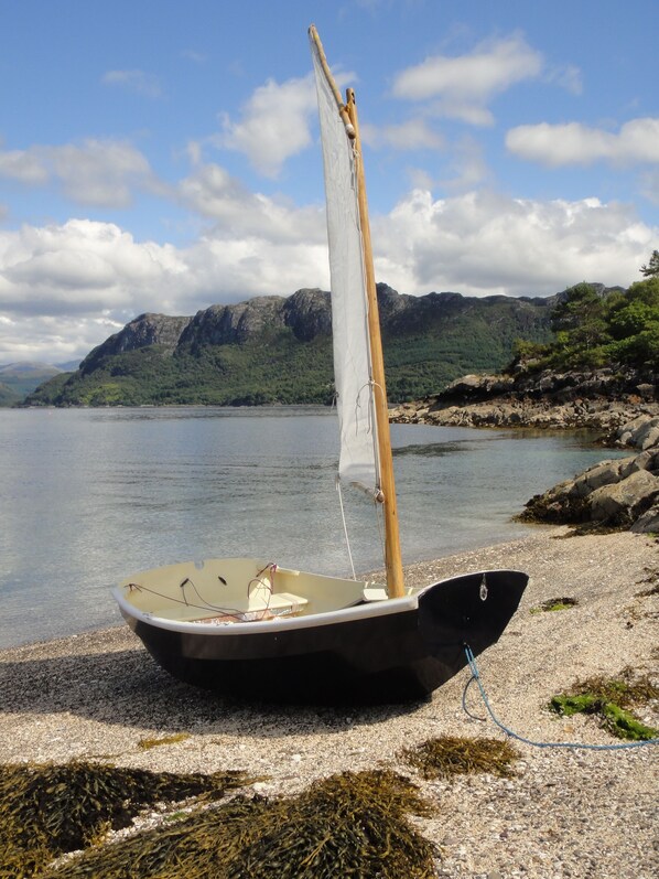 A small sailboat with a wooden mast and white sail, docked on a pebble beach with rocky outcrops and mountains in the background. - Ratagan Youth Hostel (Kyle)