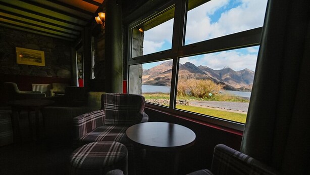 A cozy room with plaid furniture, a round table, and a large window offering a view of mountains and a lake. - Ratagan Youth Hostel (Kyle)