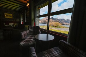 A cozy room with plaid furniture, a round table, and a large window offering a view of mountains and a lake. - Ratagan Youth Hostel (Kyle)