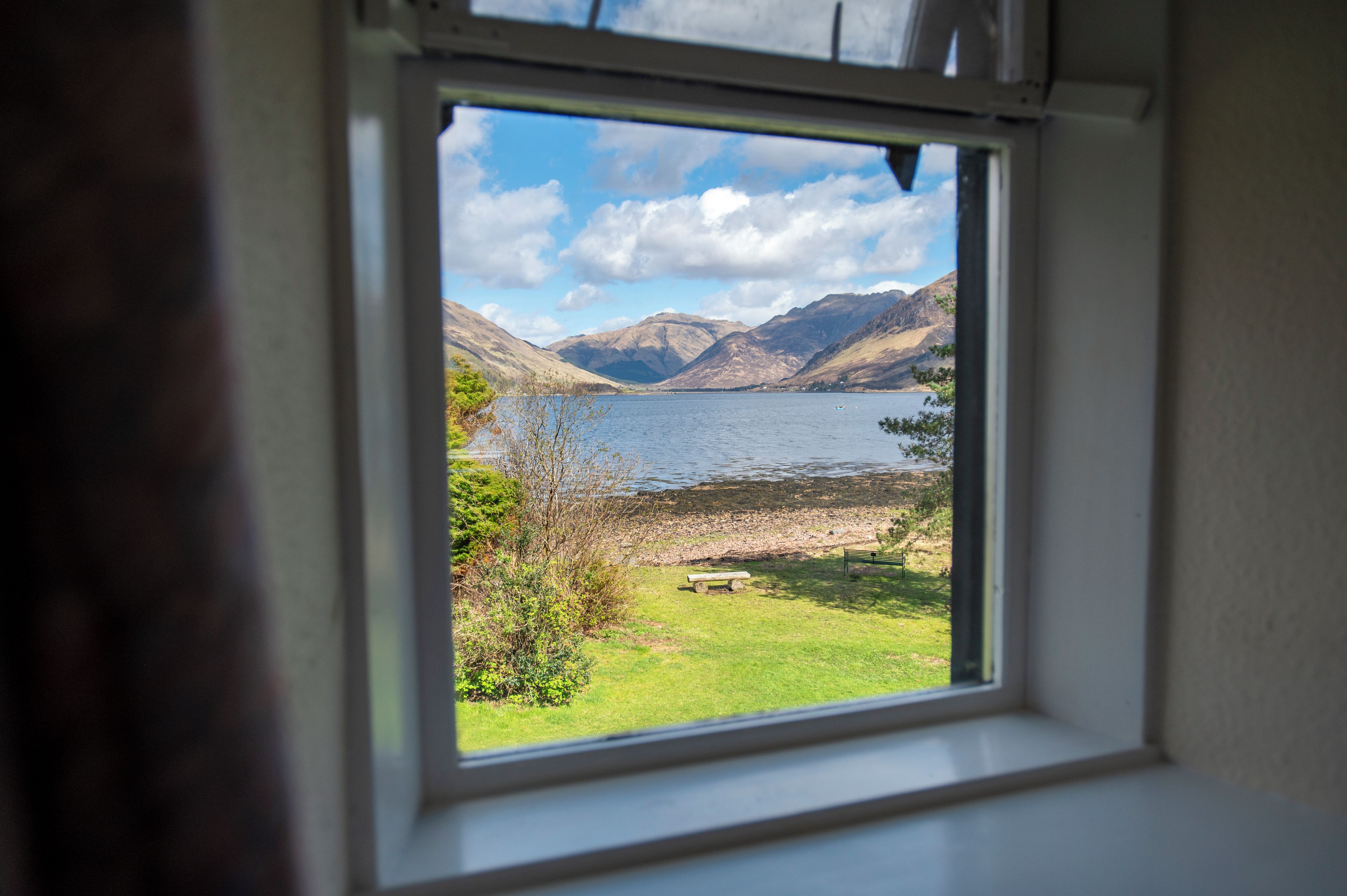 A window with a view of a lake, mountains, and a grassy area with a bench.