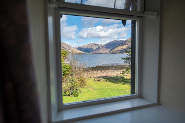 A window with a view of a lake, mountains, and a grassy area with a bench.