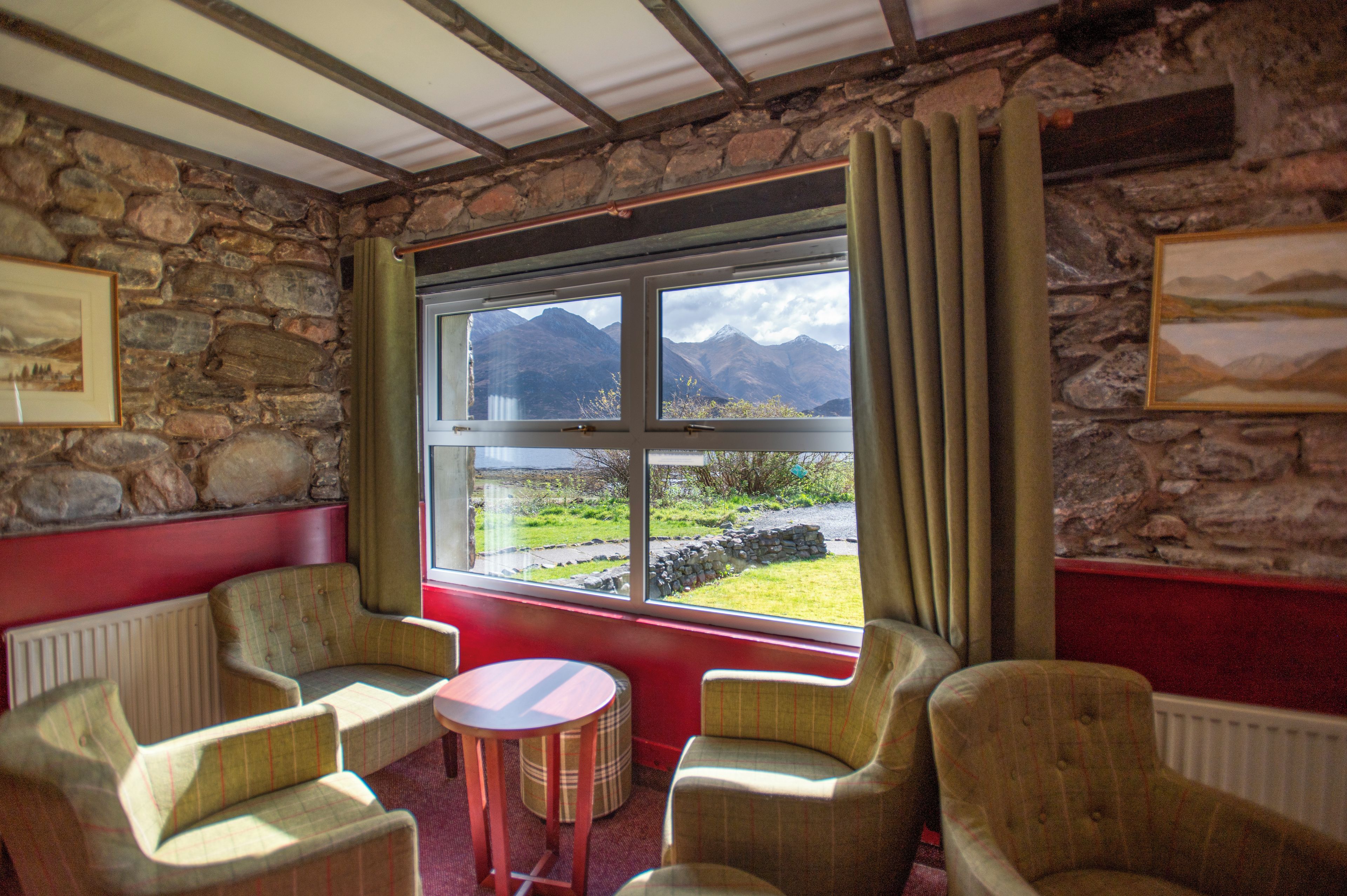 Una habitación con paredes de piedra, un ventanal con vistas a montañas y dos sillones con una mesita redonda.
