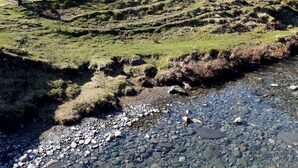 A large building with multiple windows and a sloped roof, surrounded by a grassy area and a stream. - Glenbrittle Youth Hostel (Minginish)