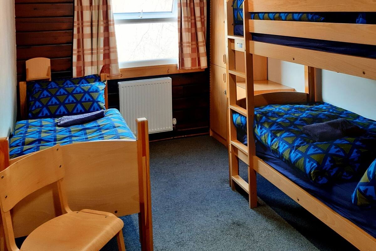 A cabin with wooden walls and a sloped ceiling. There are two bunk beds with blue patterned bedding and a single chair. A window with curtains allows natural light into the room.